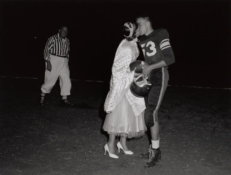 A football player in uniform kisses a woman in a formal dress and heels on a field at night, while a referee in a striped shirt watches in the background.