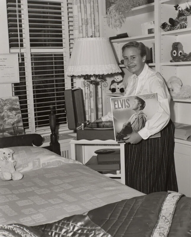 A young woman smiles while holding an Elvis Presley vinyl record in a bedroom with a record player, a bed, and shelves filled with stuffed animals.