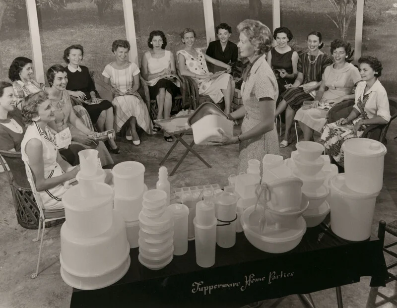 A woman demonstrates Tupperware products to a group of women seated in a semi-circle at a home party.