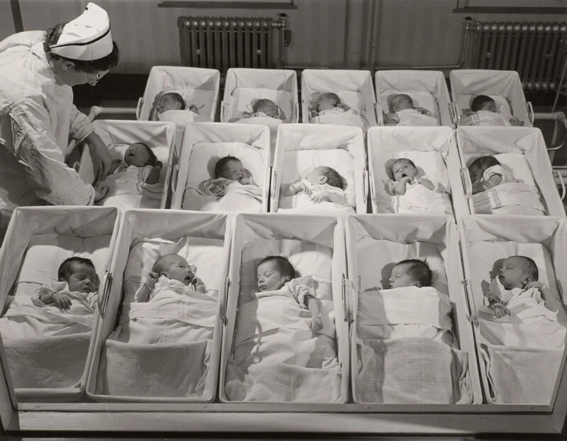 A nurse in a white uniform and cap tends to a row of newborns sleeping in individual hospital bassinets.