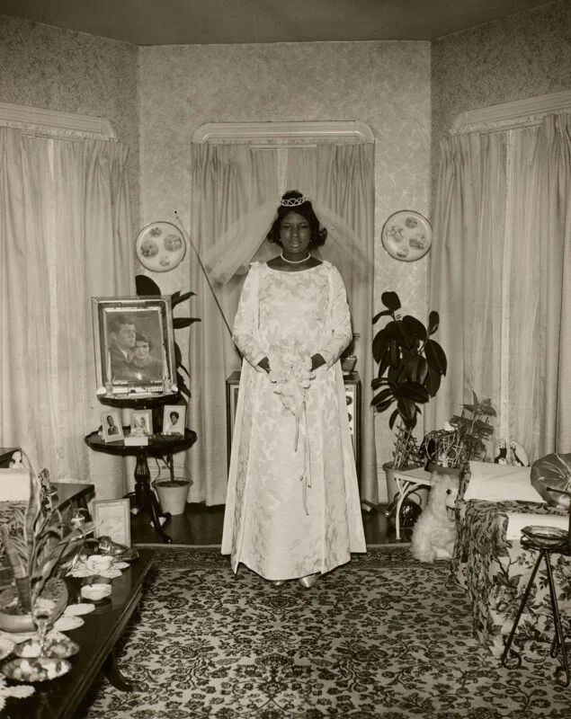 A Black bride in a long-sleeved white wedding dress and veil stands in a living room, with a framed portrait of John and Jackie Kennedy on a side table nearby.