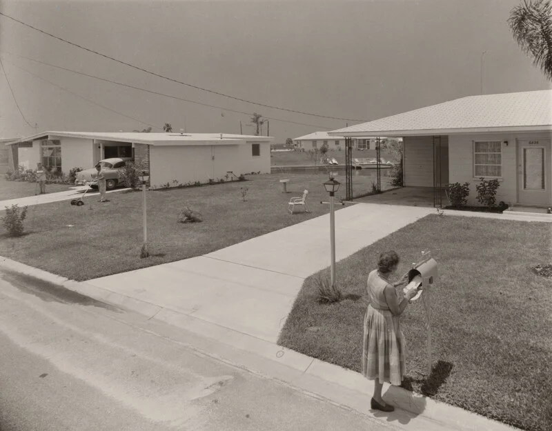 A woman in a dress checks her mailbox in a suburban neighborhood while a man mows the lawn of a nearby house.