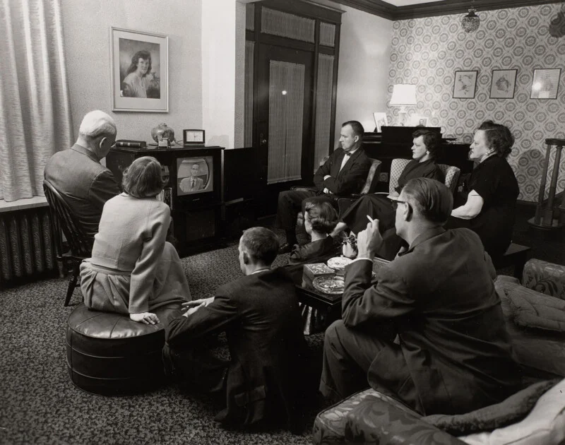 A group of seven people sit in a living room watching a man on a television set.