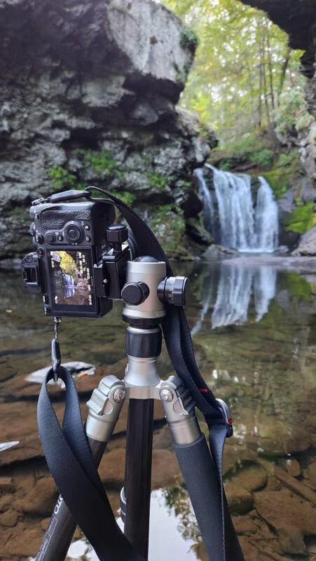 A camera mounted on a tripod stands in a shallow stream in front of a waterfall, with the camera's screen displaying a preview of the scene.