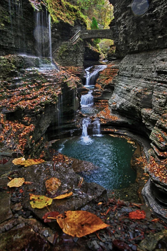 A stone bridge arches over a tiered waterfall cascading into a deep, dark pool surrounded by layered rock cliffs and autumn leaves.