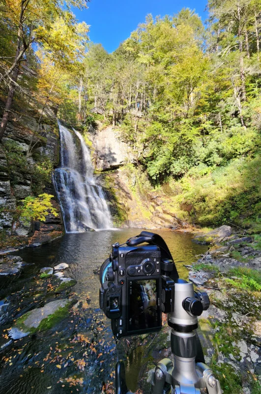 A camera on a tripod faces a waterfall cascading into a pool surrounded by rocky, forested terrain.