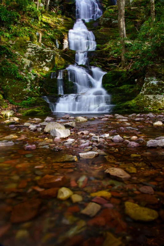 A multi-tiered waterfall cascades down a mossy, rocky cliff into a shallow, clear stream filled with colorful pebbles.