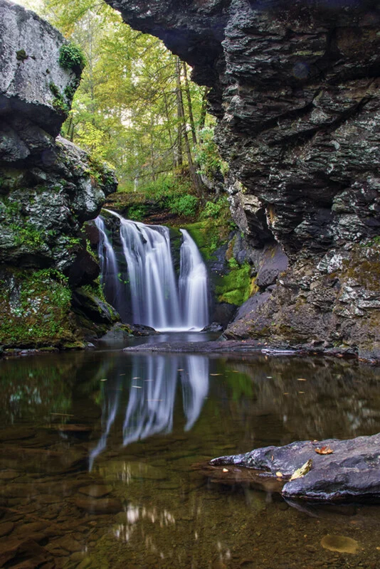 A waterfall cascades between two large, mossy rock formations into a calm pool reflecting the water and surrounding trees.