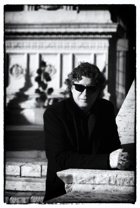 A man with curly hair and sunglasses wearing a dark jacket leans against a stone ledge in front of a classical building.