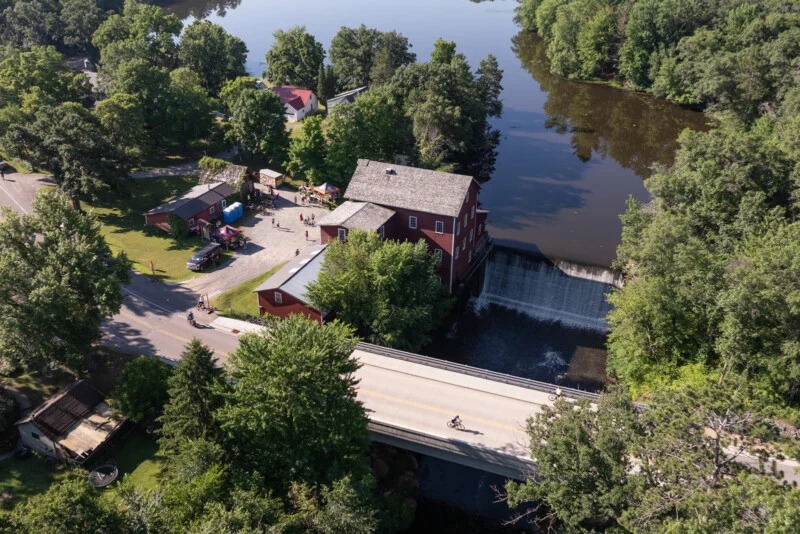 An aerial view shows a red mill building next to a waterfall and a bridge where cyclists are riding over a river.