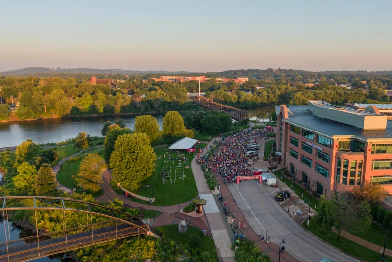 An aerial view shows a large crowd of cyclists gathered at a starting line on a street next to a river, a park, and a multi-story brick building.