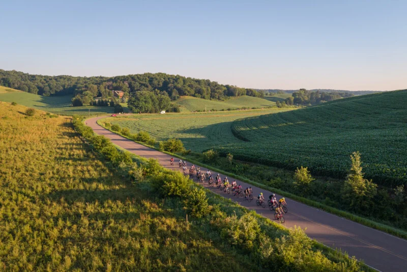 A large group of cyclists rides in a line along a winding paved road through a rural landscape of rolling green hills and fields.