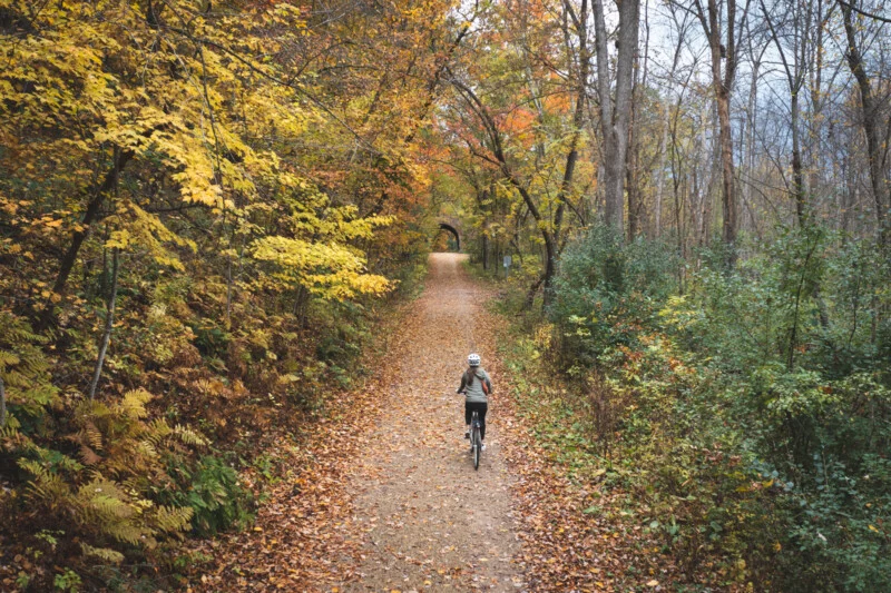 A person wearing a helmet rides a bicycle away from the camera along a leaf-covered trail through an autumn forest toward a distant stone tunnel.