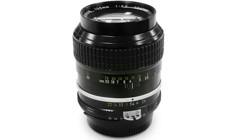 A black Nikon Nikkor 105mm f/2.5 manual focus camera lens standing upright against a white background.