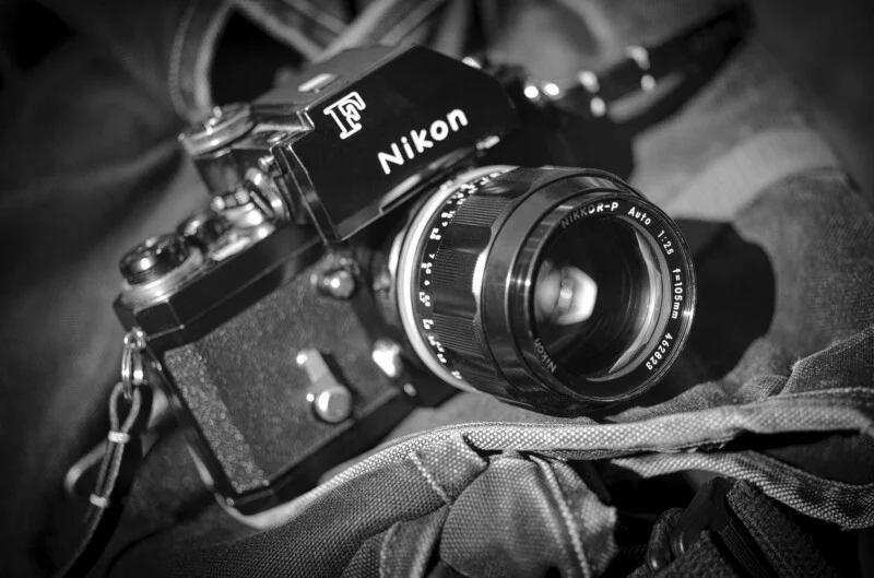 A black and white close-up of a vintage Nikon F camera with a Nikkor-P 105mm lens resting on a textured fabric surface.