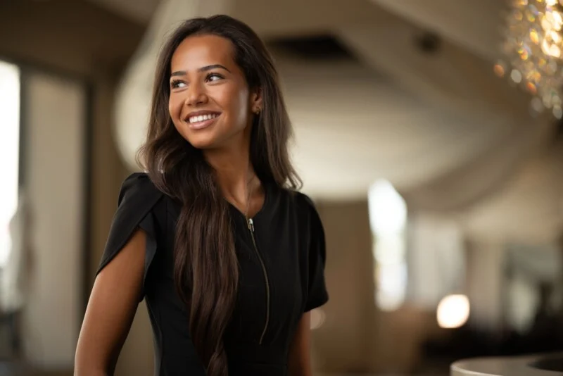 A smiling woman with long, dark hair wears a black dress with a front zipper, looking off to the side in a softly lit room.