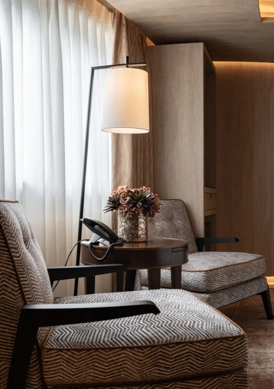 Two patterned armchairs face a small round wooden table holding a telephone and a vase of flowers, illuminated by a floor lamp.