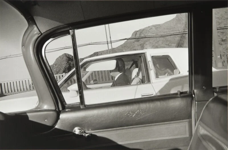 A view from inside a car looking out the window at a white car driving alongside, with a person visible in the driver's seat.