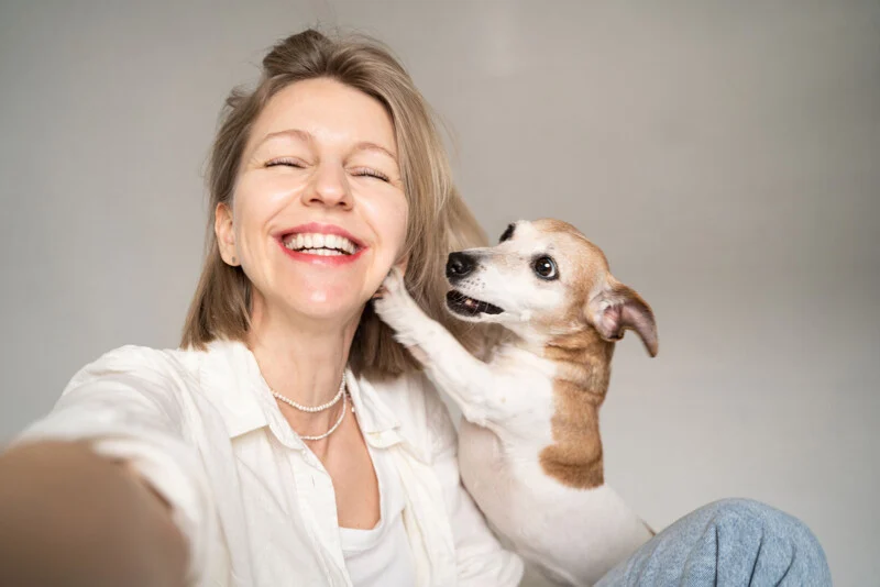 A smiling woman with blonde hair takes a selfie while a small Jack Russell Terrier touches her cheek with its paw.