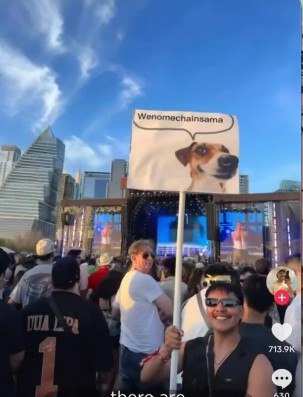 A person holds a sign featuring a dog and the text "Wenomechainsama" at an outdoor concert.