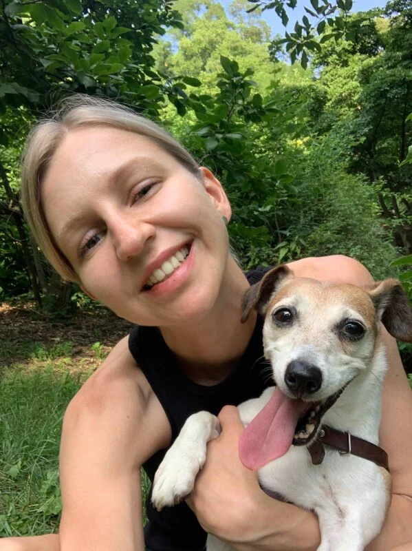 A smiling woman with blonde hair holds a small, white and brown Jack Russell Terrier with its tongue hanging out in a park.