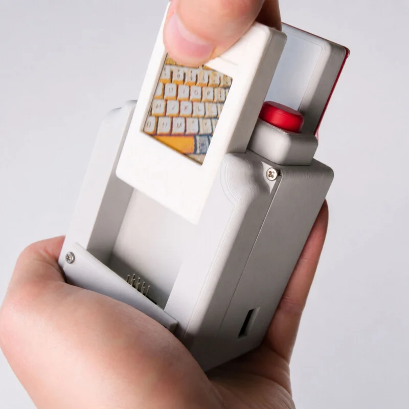 A hand holds a 3D-printed gray device while inserting a white slide containing a photograph of a keyboard into its top slot.