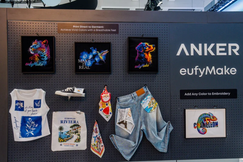 A display board features samples of direct-to-garment printing and embroidery, including framed artwork, a t-shirt, a tote bag, a shoe, scarves, and denim shorts, with the brand name "ANKER eufyMake" visible.