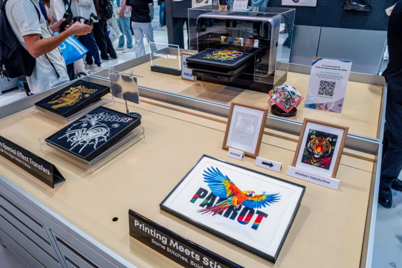A display table features a fabric printer, framed embroidered samples, and printed textile art, including a parrot design.