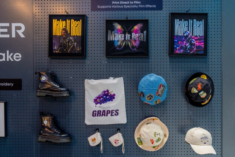 A pegboard display features three framed "Make It Real" prints, two black boots, a white tote bag with a grape graphic, two small pouches, two bucket hats, and a baseball cap.