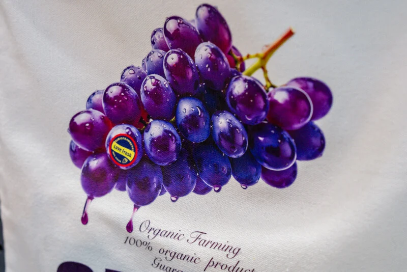 A bunch of purple grapes with water droplets on a white fabric background, featuring a sticker that reads "Love Fresh" and text below stating "Organic Farming 100% organic product".