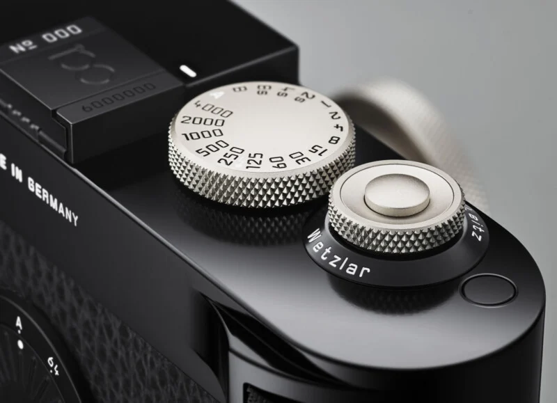 Close-up of the silver dials on a black Leica camera, showing details like shutter speed settings and the engraved word "Wetzlar" on one dial, with textured metal surfaces and part of the camera body visible.
