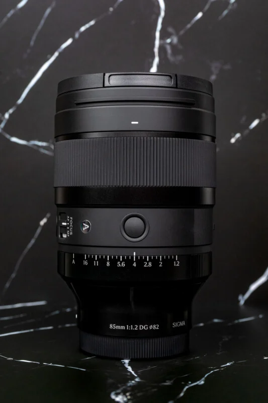 A black Sigma 85mm 1:1.2 DG Ø82 camera lens stands upright against a black and white marble-patterned background.