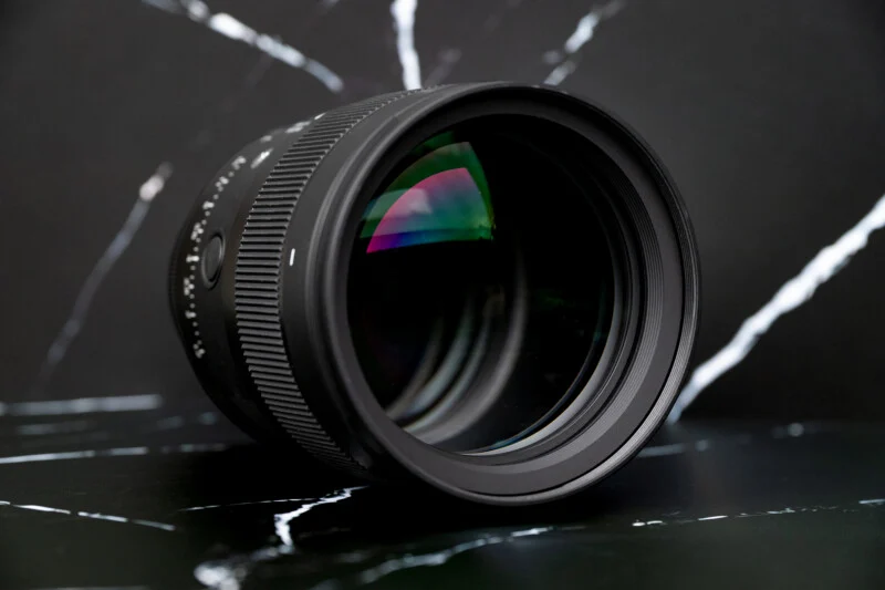 A black camera lens sits on a dark, white-veined marble surface against a matching black marble background.