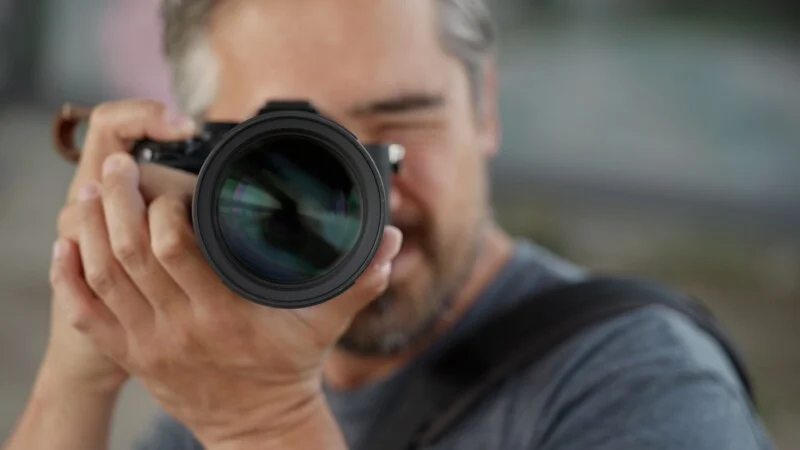 A man with gray hair holds a camera up to his eye, focusing the lens toward the viewer.
