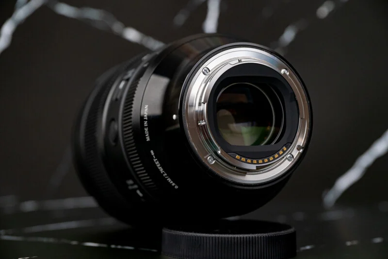 A black camera lens is positioned on a dark, marble-patterned surface, showing its rear mount, gold electronic contacts, and "MADE IN JAPAN" text.