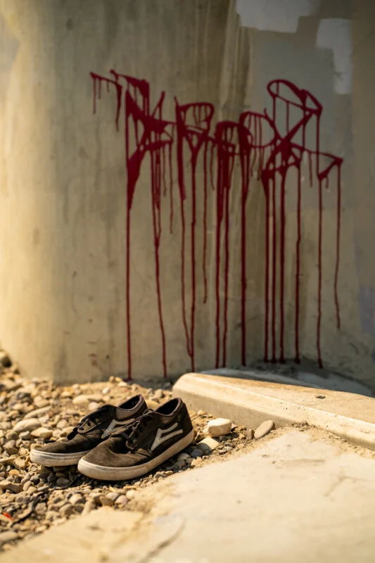 A pair of worn, dark-colored skate shoes sits on a gravel surface in front of a concrete wall marked with red graffiti drips.
