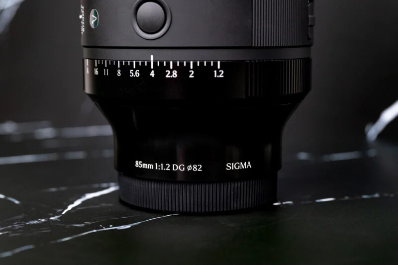 A close-up of a black camera lens standing upright on a black marble surface, showing aperture markings from 1.2 to 16 and text reading 85mm 1:1.2 DG 82 SIGMA.