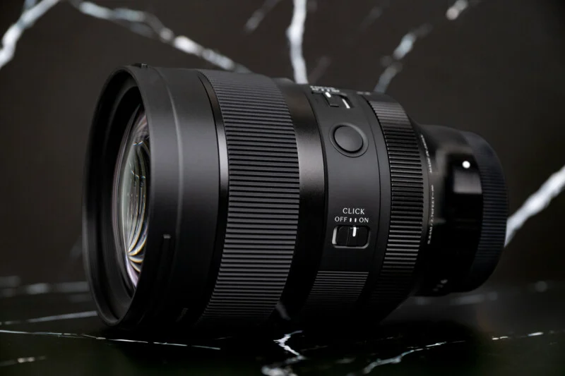A black camera lens sits on a dark, marble-patterned surface, featuring a focus ring, a customizable button, and a switch labeled "CLICK OFF | ON."
