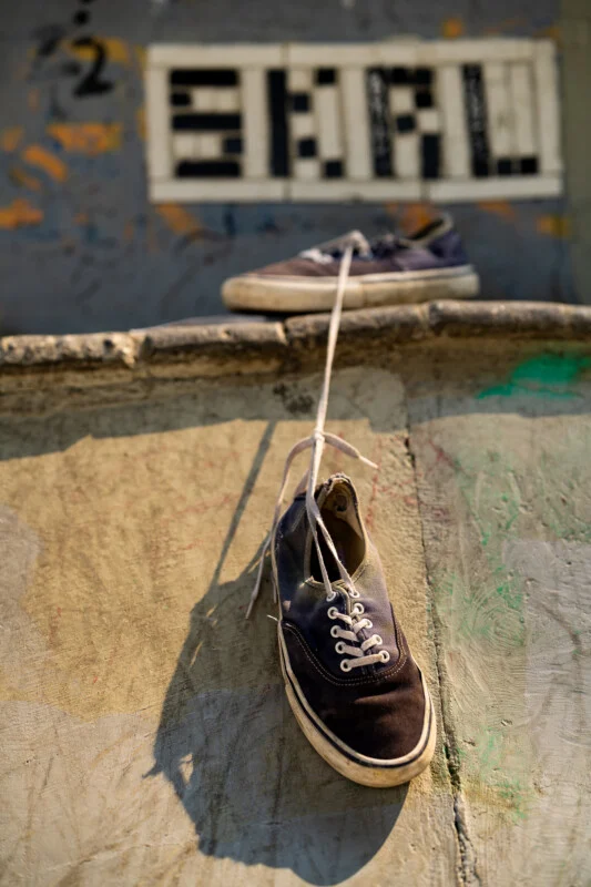 A pair of dark blue canvas sneakers are tied together by their laces, with one resting on a ledge and the other hanging down against a textured, graffiti-covered wall.