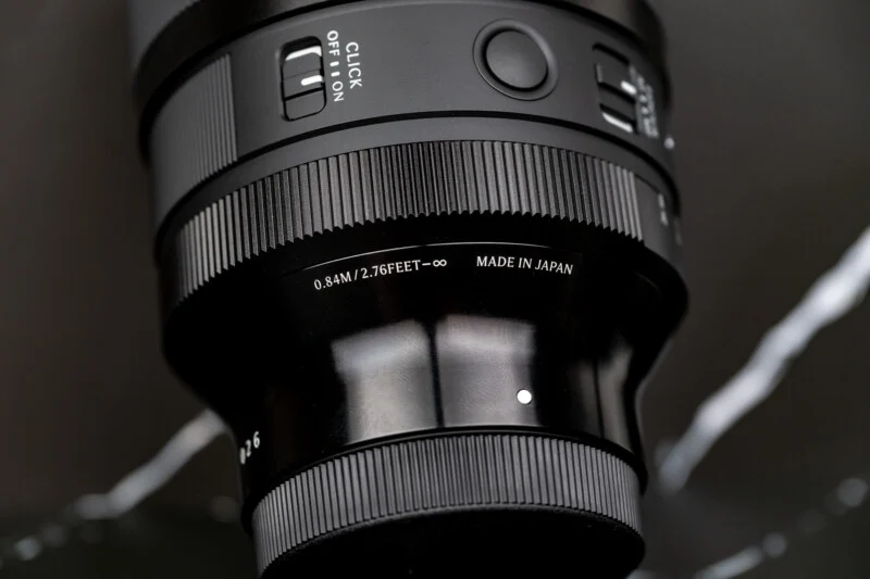 A close-up view of a black camera lens barrel featuring a focus ring, a click switch, and text indicating a minimum focus distance of 0.84M/2.76FEET and "MADE IN JAPAN."