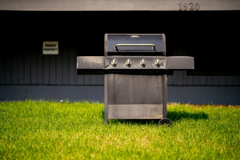 A black gas grill stands on a green lawn in front of a gray building with the number 1920 and a small sign visible on the wall.