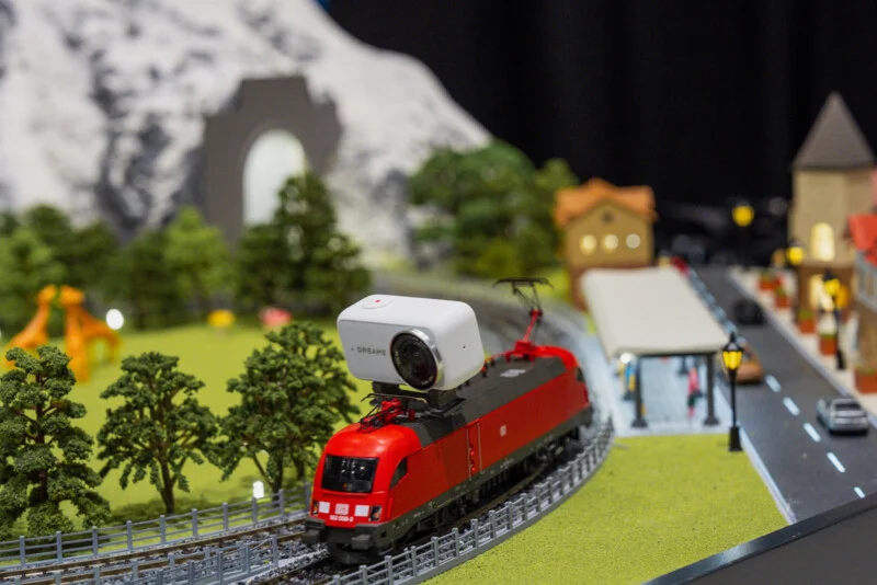 A red model train with a white camera mounted on top travels along tracks through a miniature landscape with trees, a tunnel, and a small town.