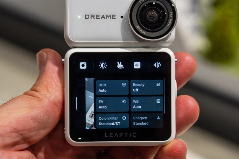A hand holds a white, square-shaped camera with a digital screen displaying a settings menu featuring options for HDR, Beauty, EV, WB, Color/Filter, and Sharpen.