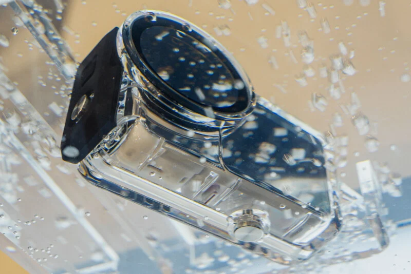 A waterproof action camera in a clear protective housing is submerged in water with visible air bubbles.