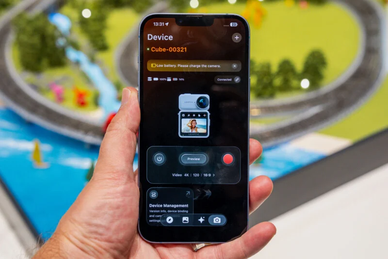 A hand holds a smartphone displaying a camera control app for a device named Cube-00321, showing a low battery warning and a live preview of a person on the screen.