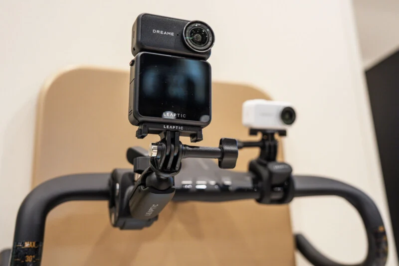 Two cameras are mounted on the handlebars of a stationary exercise bike, with the larger camera in the foreground displaying the brand name LEAPTIC.
