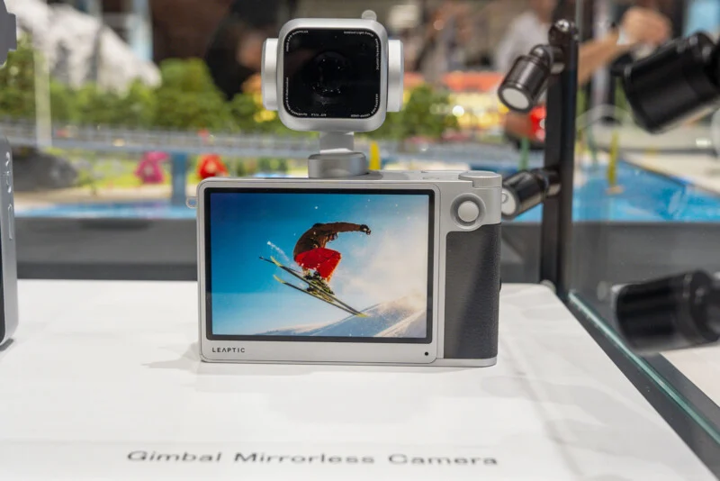 A silver LEAPTIC gimbal mirrorless camera sits on a white display surface, showing a skier on its screen.