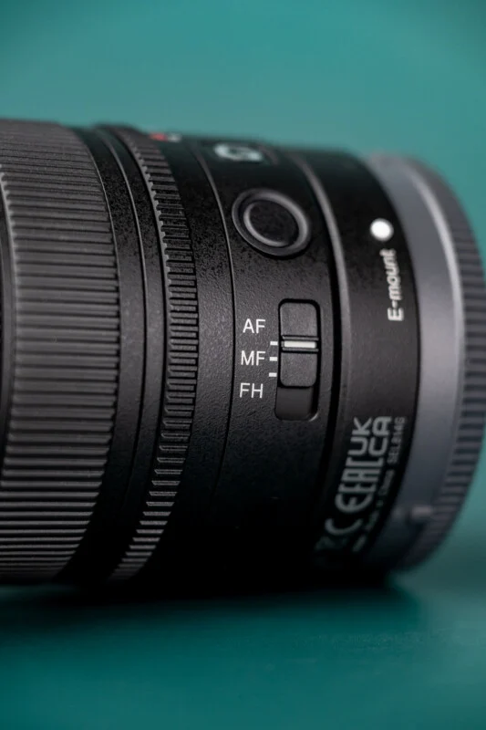 A close-up of a camera lens barrel featuring an E-mount label and a focus mode switch set to MF, positioned against a teal background.