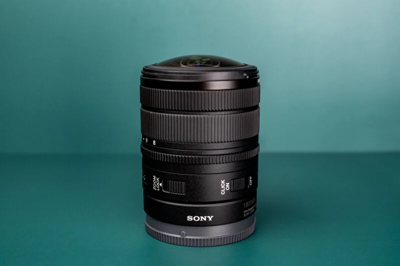 A black Sony camera lens stands upright on a teal surface against a matching teal background.