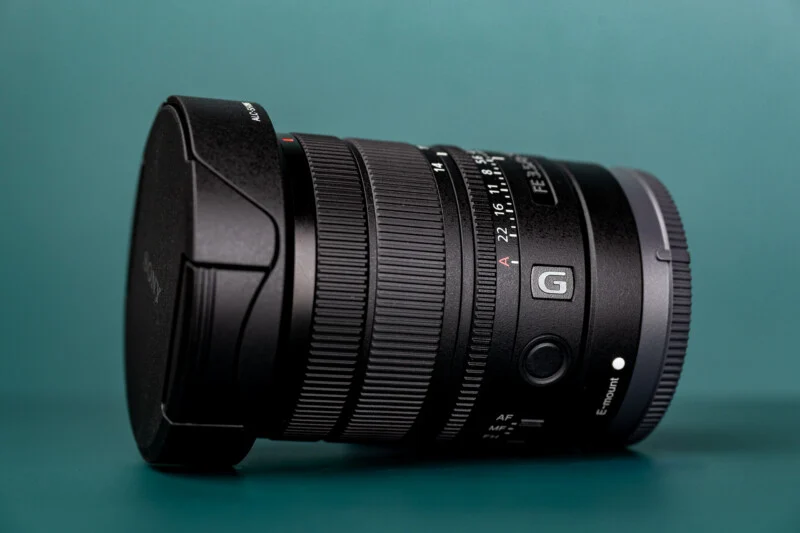 A black Sony G-series camera lens with a lens cap attached, resting on a teal surface.
